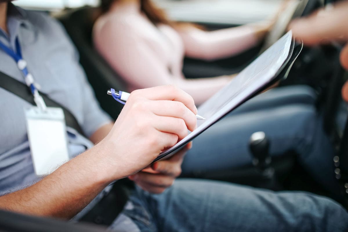 Examiner's hand marking a clipboard form on his lap, ID lanyard visible, learner's hands on the wheel beside him