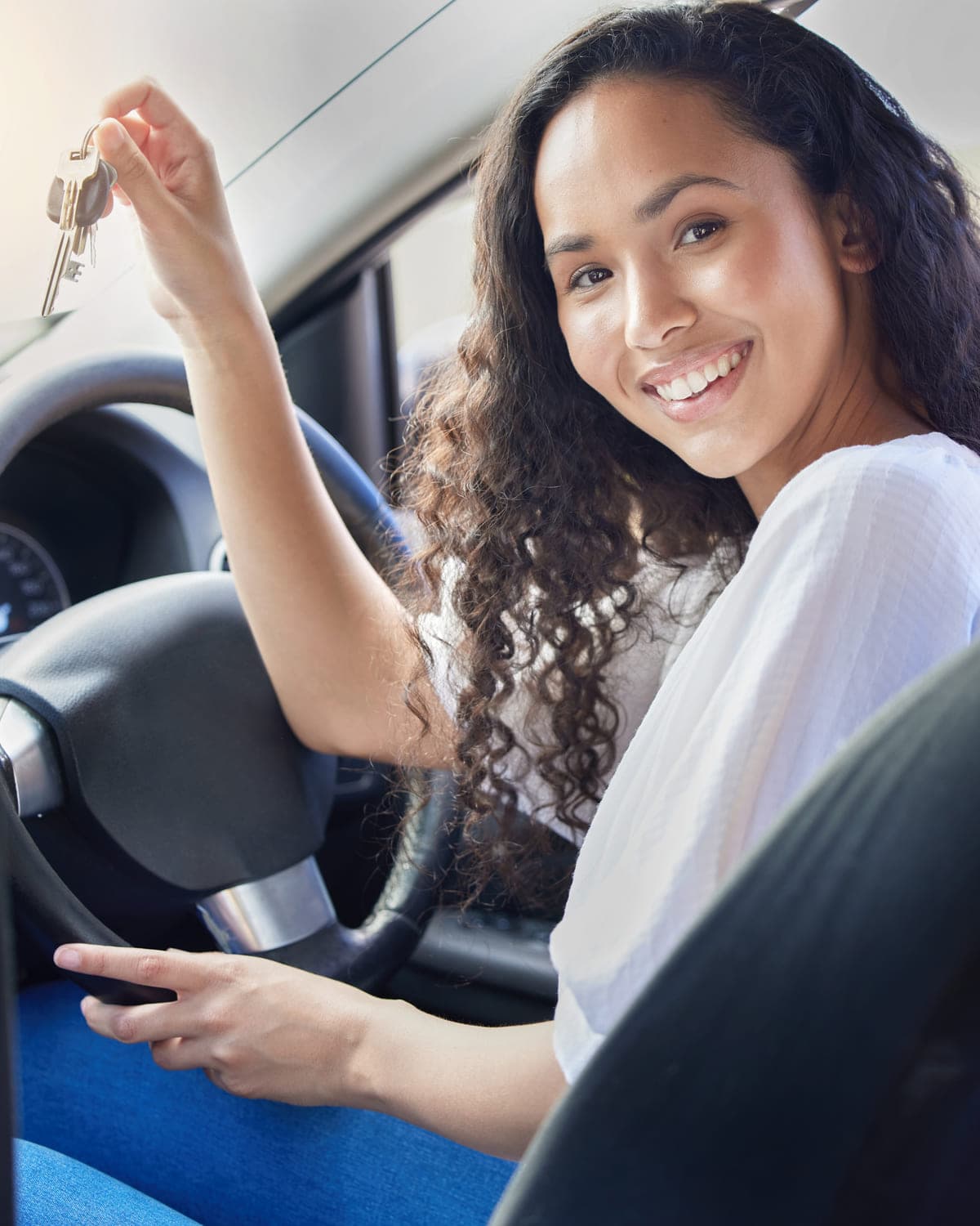 Young woman in the driver's seat, one hand on the steering wheel, holding up a set of car keys and smiling